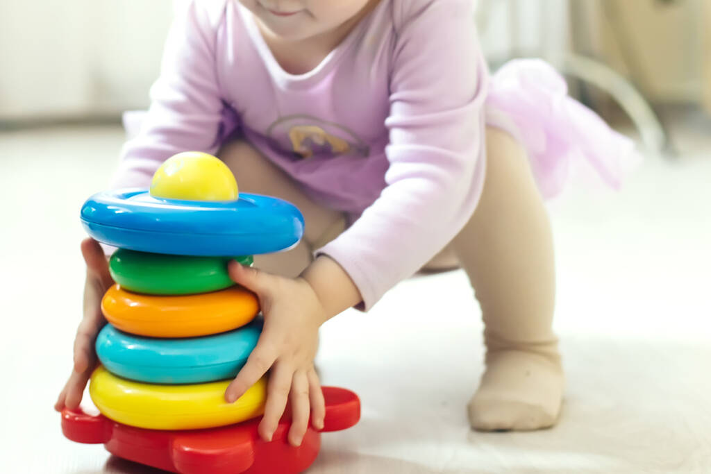 girl plays logical pyramid on floor in living room on sunny day. Montessori wooden toy folded pyramid. Circle, quadra, triangle, rectangle wooden elements of children's toys. Multi-colored toy blue, yellow, red, green
