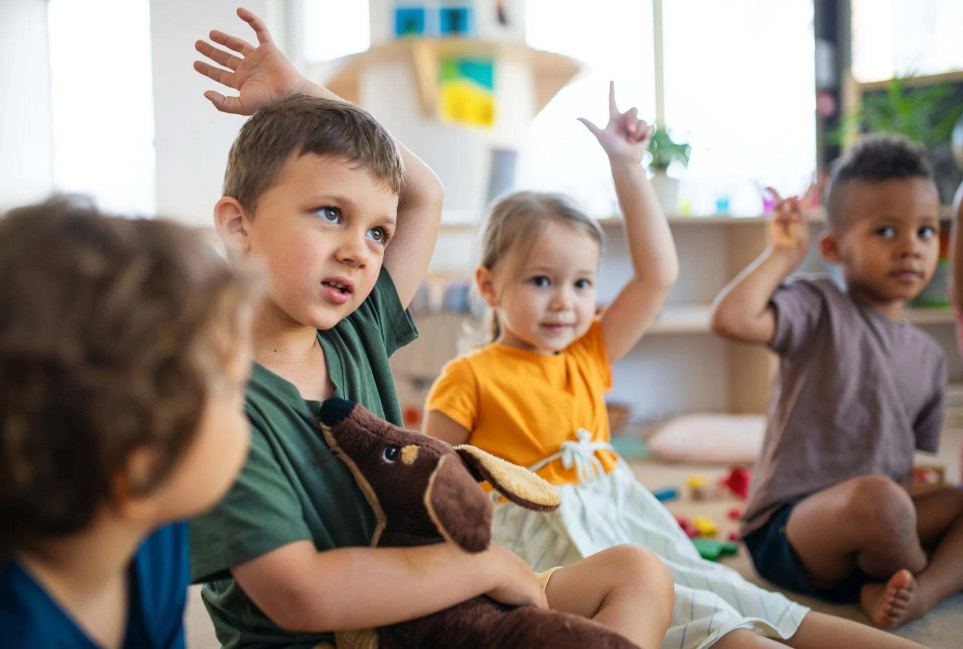 group-of-small-nursery-school-children-sitting-on-floor-indoors-in-classroom-raising-hands